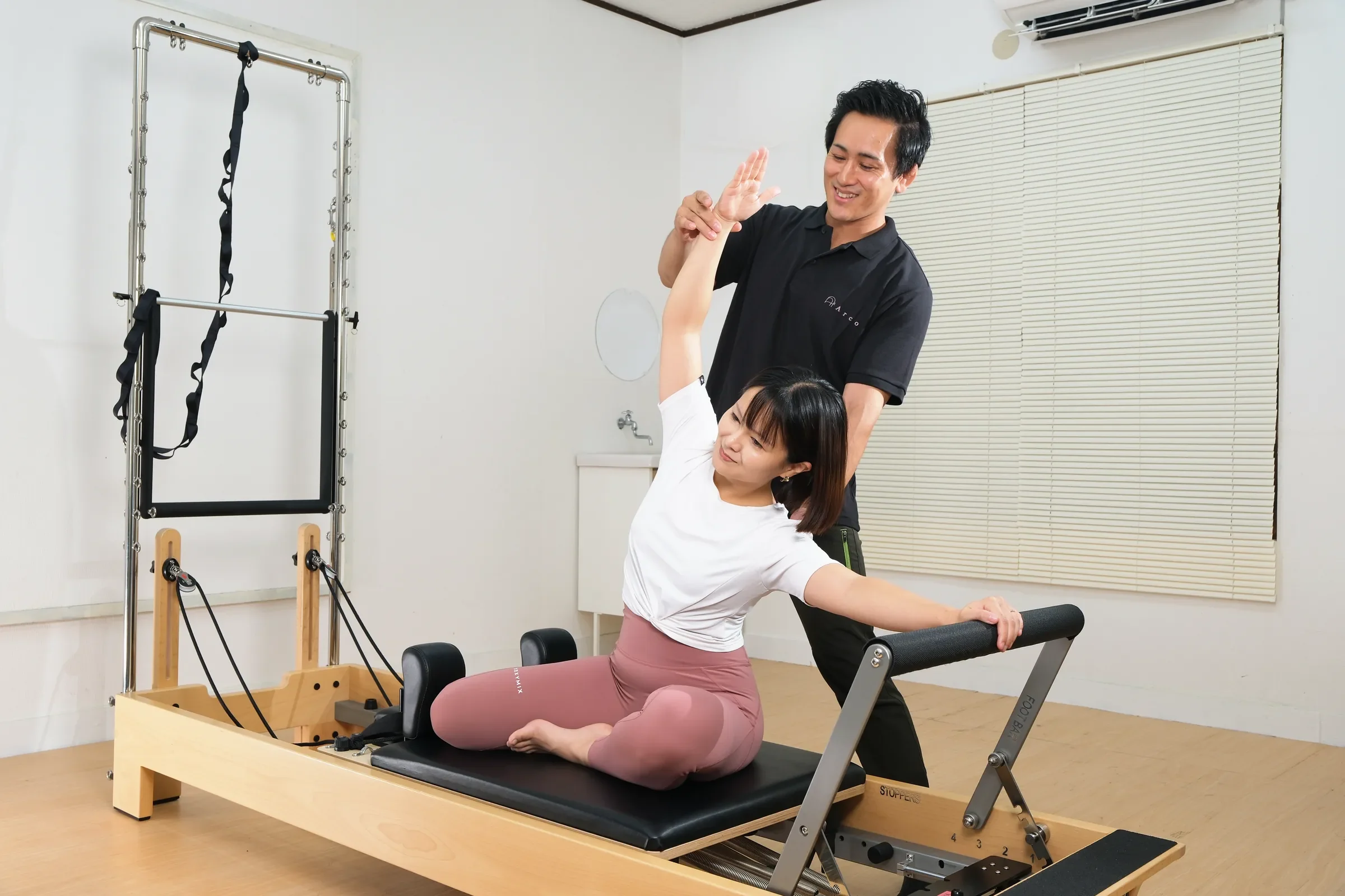 トレーナーの補助を受けながら、リフォーマーに座って体側を伸ばす女性