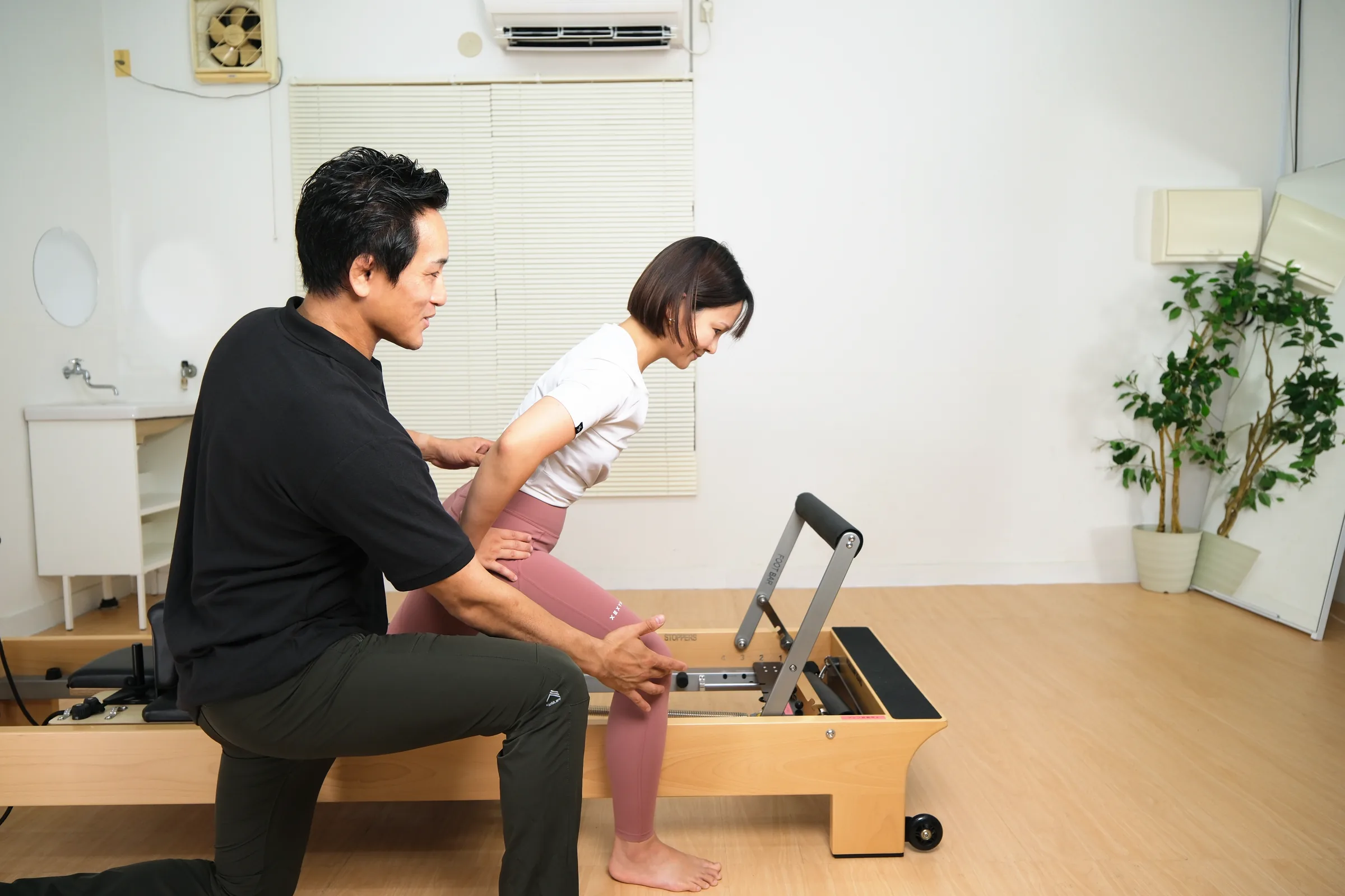しゃがんだ姿勢で脚の動きを確認しながら、リフォーマーでのエクササイズをサポートするトレーナー