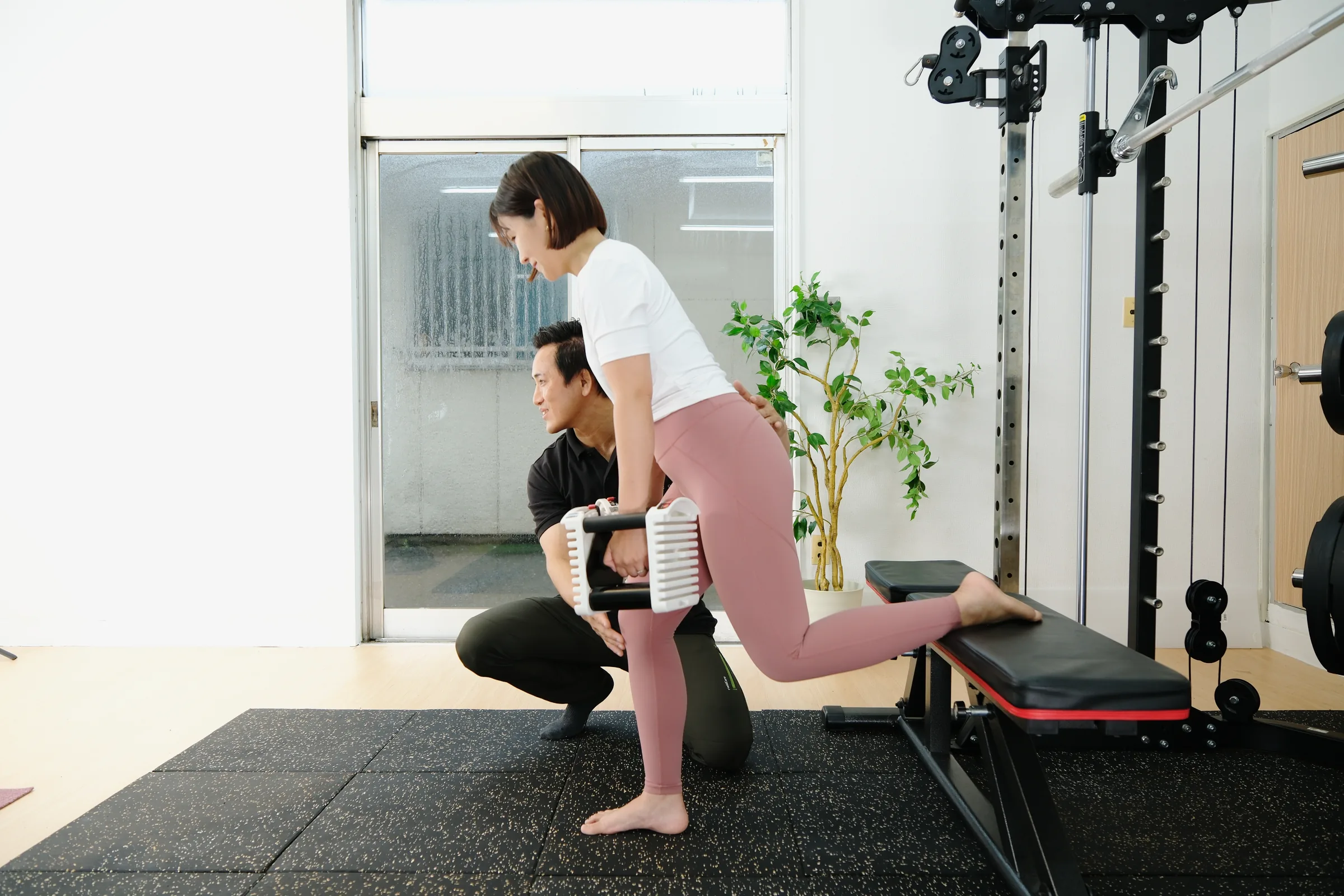 ベンチに片足を乗せてダンベルを持つトレーニングを、トレーナーがしゃがんで膝の位置を確認しながら指導している様子